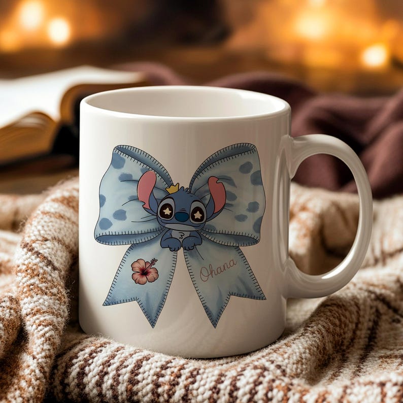 Pu&ograve; includere: Tazza in ceramica bianca con un personaggio dei cartoni animati blu con una corona, su un grande fiocco blu a pois bianchi. Un fiore di ibisco rosa e la parola "Ohana" sono anch'essi presenti. La tazza ha un manico bianco.
