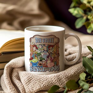 K&ouml;nnte beinhalten: Wei&szlig;e Keramiktasse mit einer farbenfrohen Grafik von Toy Story-Charakteren, dem Text "TOY STORY" und "SINCE 1995". Die Tasse enth&auml;lt ein hei&szlig;es Getr&auml;nk und steht auf einer beigefarbenen Strickdecke.