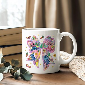 May include: White ceramic mug featuring a colorful bow design with cartoon characters. The bow is decorated with butterflies, carrots, and flowers. The mug has a curved handle and is set on a wooden surface.