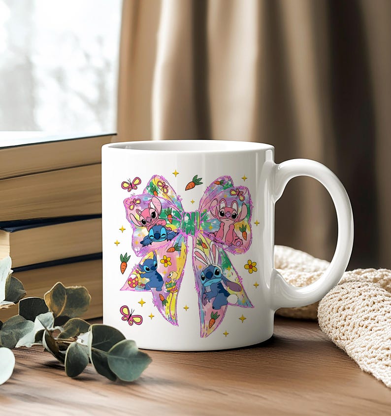 May include: White ceramic mug featuring a colorful bow design with cartoon characters. The bow is decorated with butterflies, carrots, and flowers. The mug has a curved handle and is set on a wooden surface.