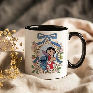 May include: White ceramic mug with a black interior and handle. The mug features a framed illustration of Lilo and Stitch, surrounded by flowers and a blue ribbon. The design is colorful and whimsical.