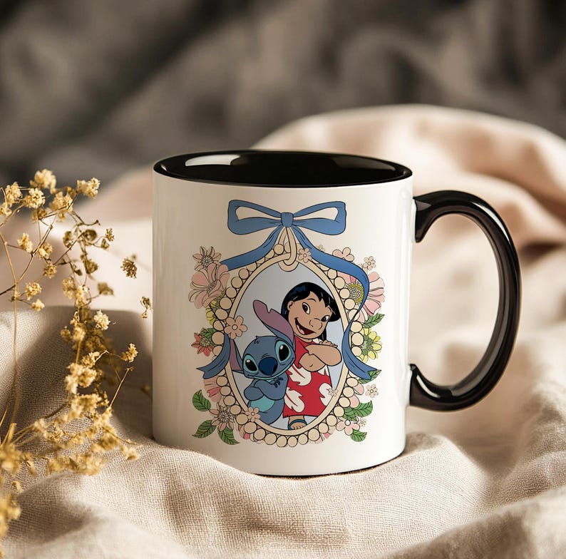 May include: White ceramic mug with a black interior and handle. The mug features a framed illustration of Lilo and Stitch, surrounded by flowers and a blue ribbon. The design is colorful and whimsical.