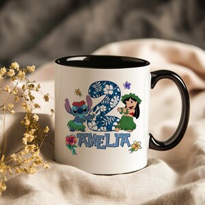 May include: White ceramic mug with a black interior and handle. The mug features a colourful design with the number 2, cartoon characters, and the name "AMELIA" in blue. Floral accents complete the design.