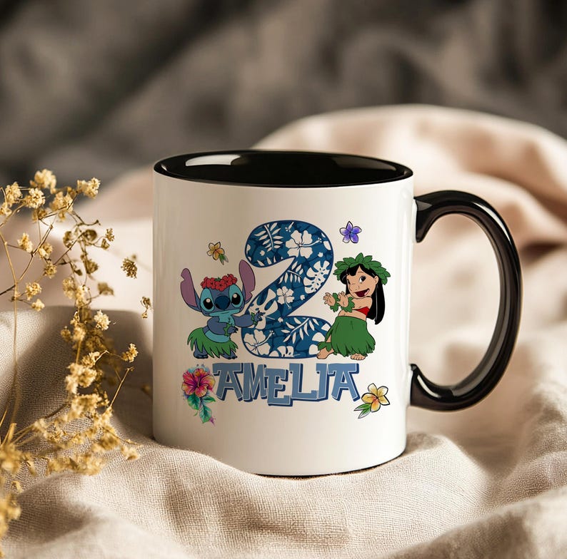 May include: White ceramic mug with a black interior and handle. The mug features a colourful design with the number 2, cartoon characters, and the name "AMELIA" in blue. Floral accents complete the design.