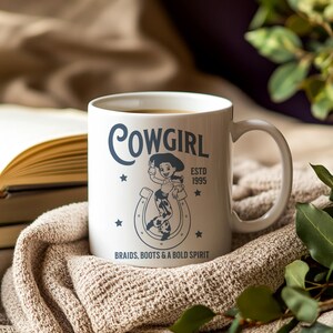 Pu&ograve; includere: Tazza in ceramica bianca con la scritta "Cowgirl" in corsivo, con una grafica di cowgirl e il testo "Braids, Boots & A Bold Spirit". La tazza ha un manico ed &egrave; riempita con una bevanda calda.