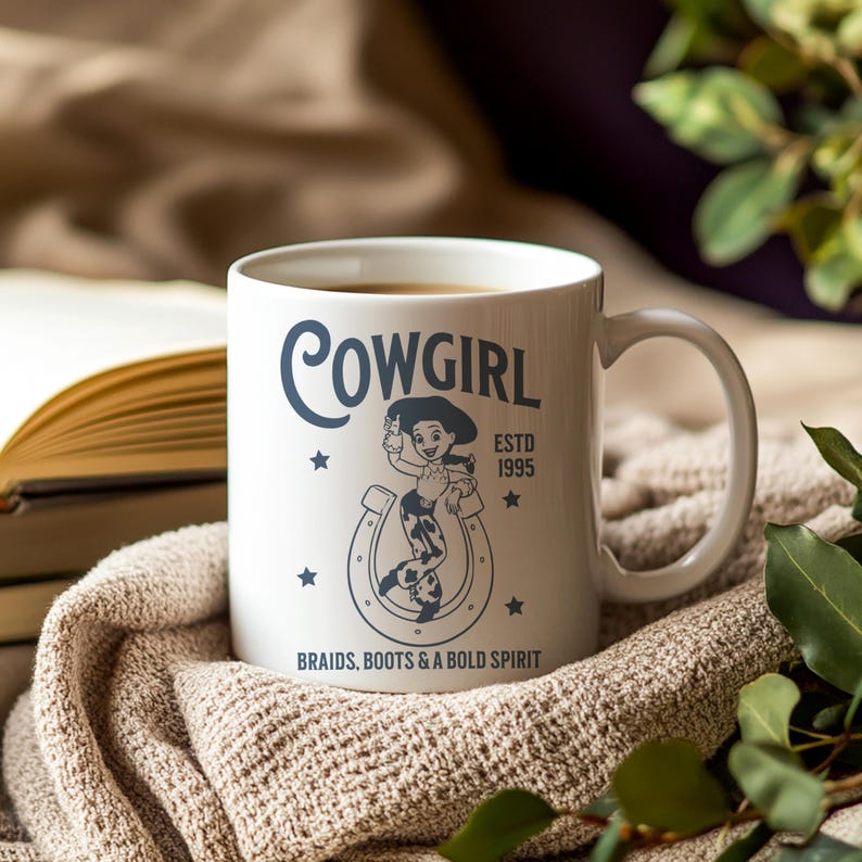 Pu&ograve; includere: Tazza in ceramica bianca con la scritta "Cowgirl" in corsivo, con una grafica di cowgirl e il testo "Braids, Boots & A Bold Spirit". La tazza ha un manico ed &egrave; riempita con una bevanda calda.