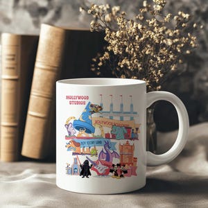 K&ouml;nnte beinhalten: Wei&szlig;er Keramikbecher mit einer farbenfrohen Illustration von Hollywood Studios Attraktionen und Charakteren. Der Becher tr&auml;gt den Text "HOLLYWOOD STUDIOS" und Bilder von ikonischen Charakteren und Wahrzeichen. Der Becher hat einen gro&szlig;en, gebogenen Griff.