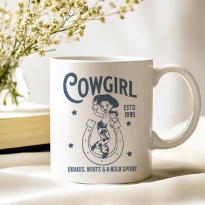 Pu&ograve; includere: Tazza in ceramica bianca con manico avvolgente con grafica cowgirl. Il design include la parola "COWGIRL", un'illustrazione di cowgirl, stelle, un ferro di cavallo e il testo "ESTD 1995 BRAIDS, BOOTS & A BOLD SPIRIT".