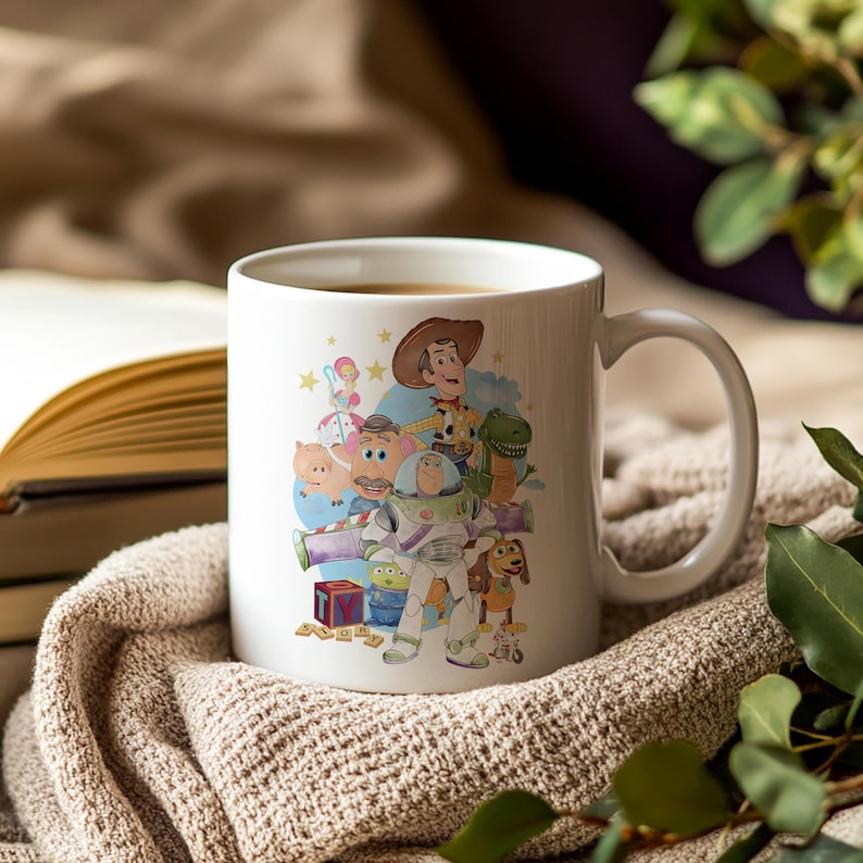 May include: White ceramic mug with a colourful illustration of characters from the animated film Toy Story. The design includes Woody, Buzz Lightyear, and other characters. The mug has a curved handle and is filled with a dark liquid.