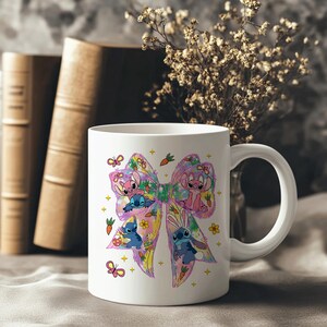 May include: White ceramic mug featuring a colorful bow design with cartoon characters, butterflies, and carrots. The bow has pink, blue, yellow, and purple accents. The mug has a white handle and is set against a neutral background with books and flowers.