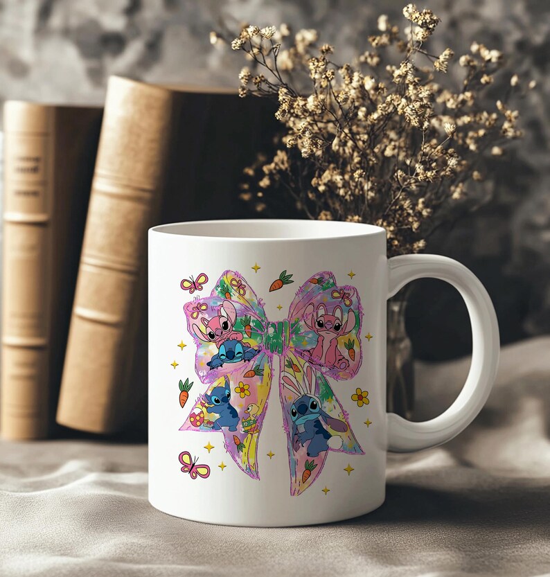 May include: White ceramic mug featuring a colorful bow design with cartoon characters, butterflies, and carrots. The bow has pink, blue, yellow, and purple accents. The mug has a white handle and is set against a neutral background with books and flowers.
