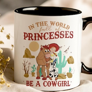May include: White ceramic mug with a black rim and handle. The mug features a cartoon image of Jessie and Bullseye from Toy Story. The text on the mug reads "In the world full of princesses be a cowgirl".