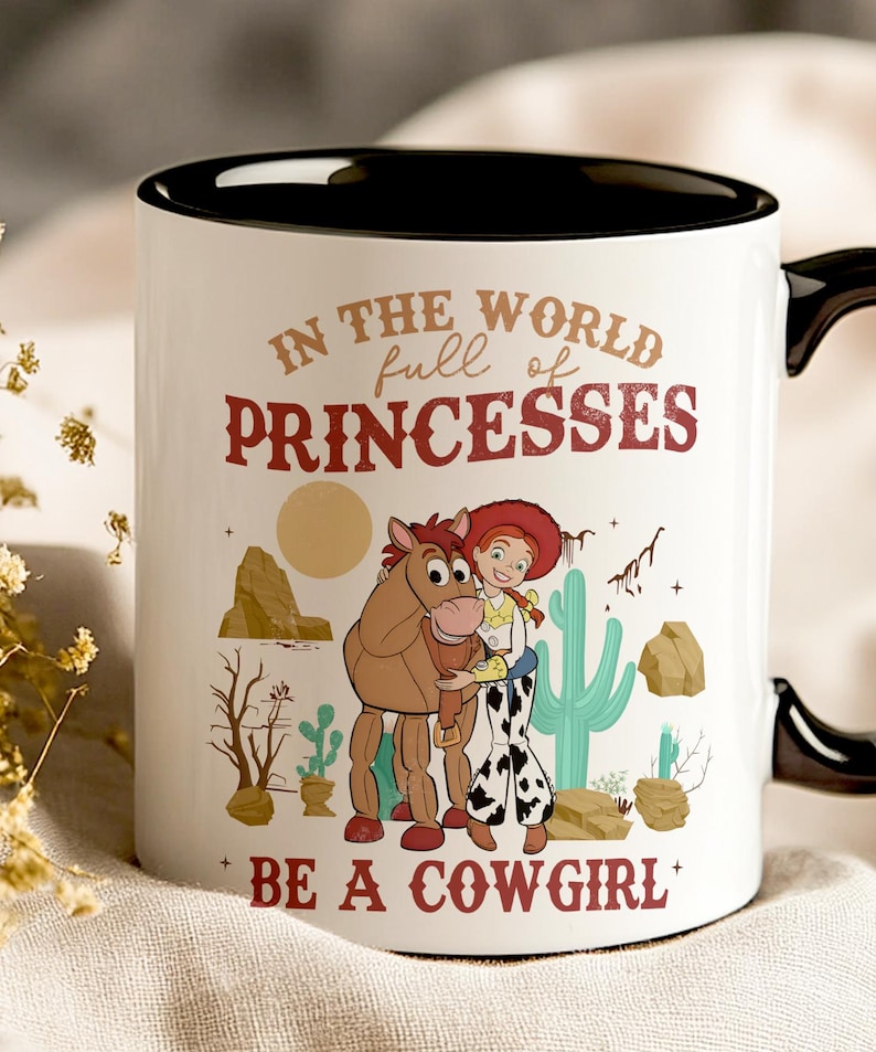 May include: White ceramic mug with a black rim and handle. The mug features a cartoon image of Jessie and Bullseye from Toy Story. The text on the mug reads "In the world full of princesses be a cowgirl".