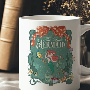 Pode incluir: Caneca de cer&acirc;mica branca com um design de "A Pequena Sereia". O design inclui Ariel, Linguado e outras criaturas marinhas, com as palavras "The Little Mermaid". Um la&ccedil;o vermelho adorna o topo do design.