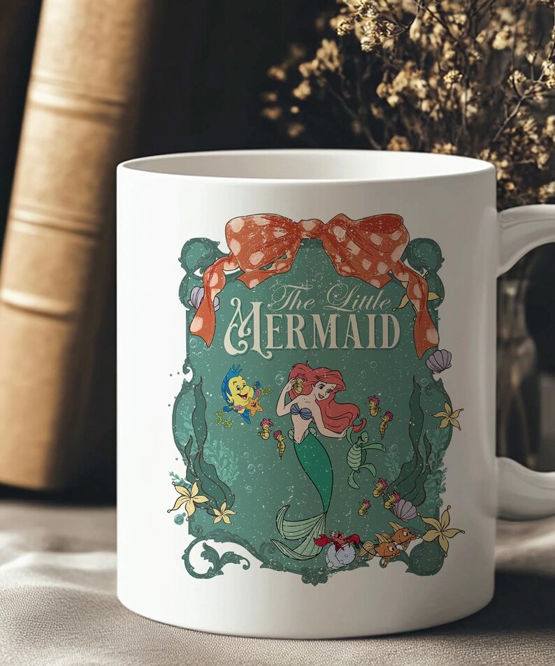 Pode incluir: Caneca de cer&acirc;mica branca com um design de "A Pequena Sereia". O design inclui Ariel, Linguado e outras criaturas marinhas, com as palavras "The Little Mermaid". Um la&ccedil;o vermelho adorna o topo do design.