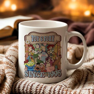 K&ouml;nnte beinhalten: Wei&szlig;e Keramiktasse mit einer farbenfrohen Grafik von Toy Story-Charakteren. Die Tasse tr&auml;gt den Text "TOY STORY" &uuml;ber den Charakteren und "SINCE 1995" darunter. Die Tasse hat einen gebogenen Griff.