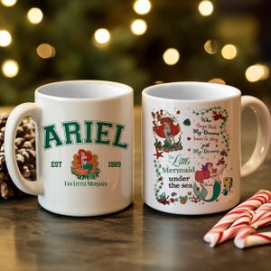 May include: Two white ceramic mugs featuring The Little Mermaid. One mug has "ARIEL" in green, "EST 1989", and a mermaid graphic. The other mug has a mermaid graphic and the text "Who Says that My Dreams have to stay just My Dreams".