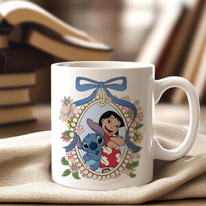 May include: White ceramic mug featuring a colorful illustration of Lilo and Stitch. The design is framed by a blue ribbon and floral accents. The mug has a curved handle and is set against a neutral background.