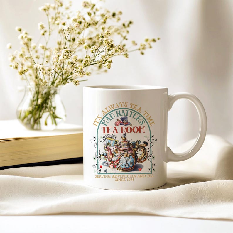 Puede incluir: Taza de cer&aacute;mica blanca con una ilustraci&oacute;n colorida de una tetera y el texto "It's Always Tea Time, Mad Hatter's Tea Room, Serving Adventures and Tea Since 1965." Un ramo de flores blancas est&aacute; en el fondo.