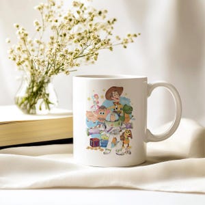 May include: White ceramic mug with a colourful illustration of Toy Story characters, including Woody and Buzz Lightyear. The mug has a curved handle and is set against a soft, neutral background. The mug is a standard size for hot beverages.