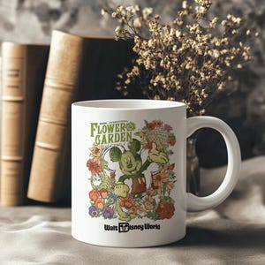 May include: White ceramic mug with a design of Mickey Mouse and flowers. The mug has the text "EPCOT INTERNATIONAL FLOWER GARDEN FESTIVAL" and "Walt Disney World". The mug is set against a neutral background with books and dried flowers.