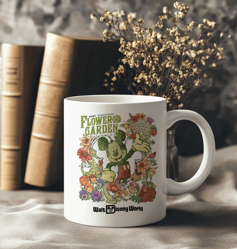 May include: White ceramic mug with a design of Mickey Mouse and flowers. The mug has the text "EPCOT INTERNATIONAL FLOWER GARDEN FESTIVAL" and "Walt Disney World". The mug is set against a neutral background with books and dried flowers.
