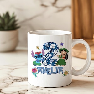 May include: White ceramic mug with a colourful design featuring the number 2, the name "AMELIA", and cartoon characters. The design includes blue, green, and floral accents. The mug has a curved handle and is set against a blurred background.