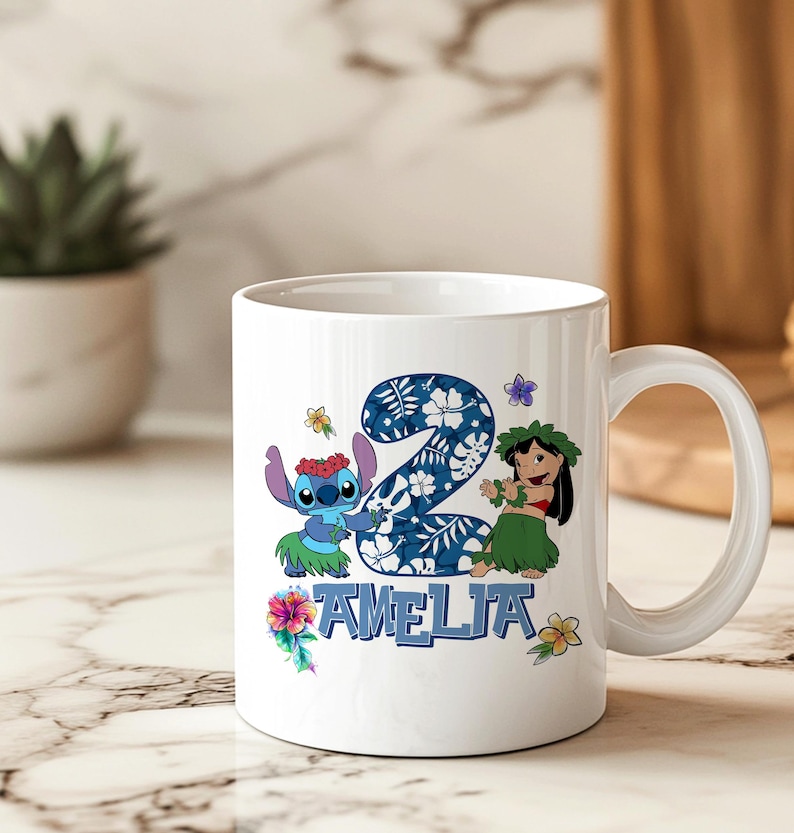 May include: White ceramic mug with a colourful design featuring the number 2, the name "AMELIA", and cartoon characters. The design includes blue, green, and floral accents. The mug has a curved handle and is set against a blurred background.