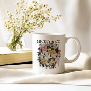 May include: White ceramic mug with a floral design featuring Mickey Mouse, Minnie Mouse, Donald Duck, and Goofy. The text "Mickey & Co. Est. 1928" is printed on the mug.