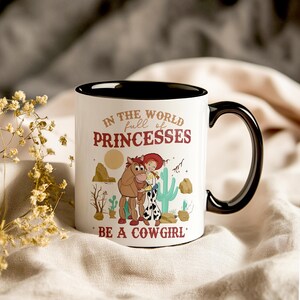 May include: White ceramic mug with a black rim and handle. The mug features a cartoon image of a cowgirl and a horse with the text "In the world full of princesses be a cowgirl".