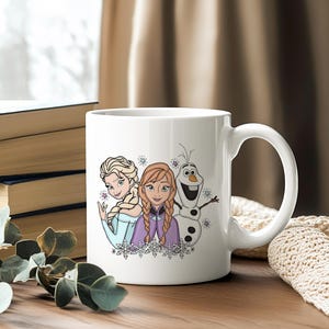 Puede incluir: Taza de cer&aacute;mica blanca con ilustraciones de Elsa, Anna y Olaf de la pel&iacute;cula Frozen. Los personajes est&aacute;n representados con sus trajes caracter&iacute;sticos, con copos de nieve y estrellas. La taza tiene un asa grande.
