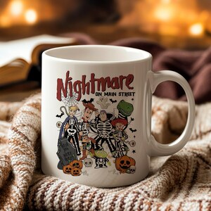May include: White ceramic mug featuring a Halloween-themed design with the text "Nightmare on Main Street." The design includes cartoon characters dressed as skeletons, pumpkins, and a tombstone. The mug has a large handle.