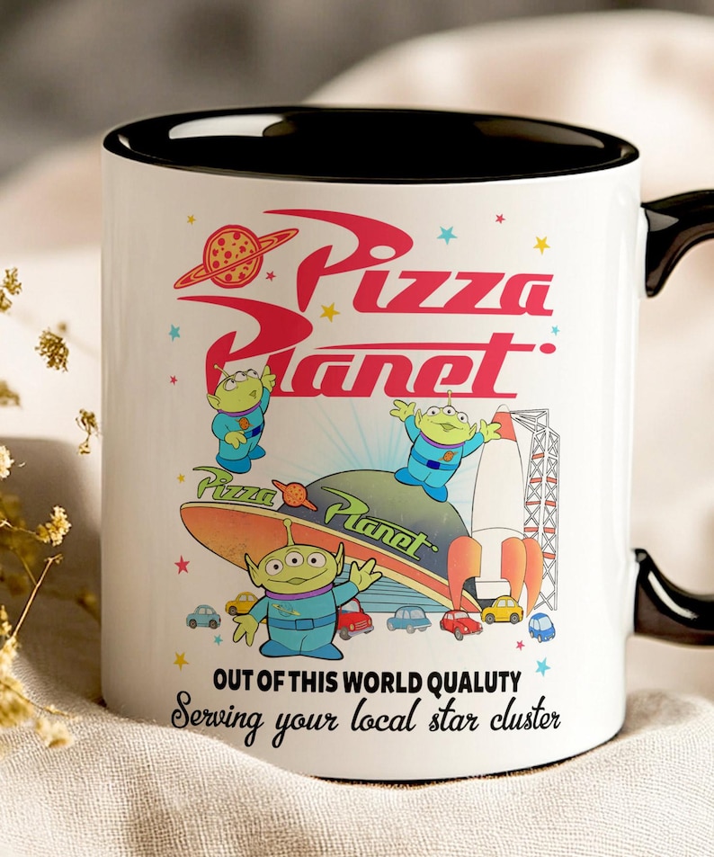 May include: White ceramic mug with a black rim and handle. The mug features a colorful illustration of the Pizza Planet restaurant from the Toy Story franchise. The text "Pizza Planet" is written in red and white with a red star and a pizza slice above the text. Three green alien characters are featured in the illustration. The text "Out of this world quality" and "Serving your local star cluster" is written in black at the bottom of the mug.
