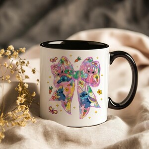 May include: White ceramic mug with a black handle and interior. The mug features a colorful bow design with cartoon characters, flowers, carrots, and butterflies. The design includes pink, blue, yellow, and green elements.