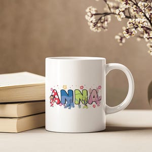 May include: White ceramic mug with the name "Anna" in colorful, cartoon-style letters. The letters are decorated with images of cartoon characters and floral accents. The mug is next to a stack of books.