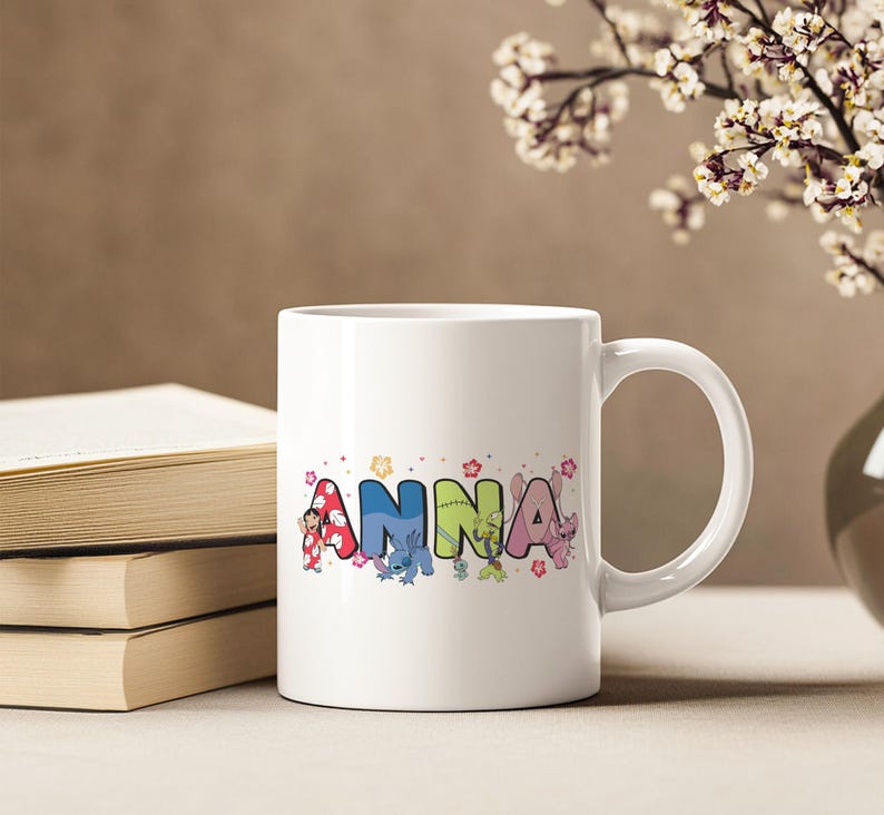 May include: White ceramic mug with the name "Anna" in colorful, cartoon-style letters. The letters are decorated with images of cartoon characters and floral accents. The mug is next to a stack of books.