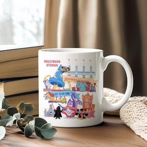 K&ouml;nnte beinhalten: Wei&szlig;e Keramiktasse mit einer farbenfrohen Illustration von Hollywood Studios-Attraktionen. Das Design enth&auml;lt den Text "Hollywood Studios" sowie verschiedene Zeichentrickfiguren und Wahrzeichen. Die Tasse hat einen gebogenen Griff und steht auf einer Holzoberfl&auml;che.