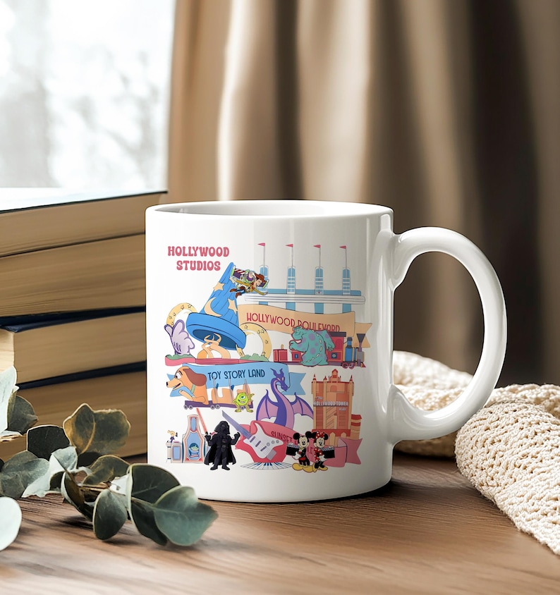 K&ouml;nnte beinhalten: Wei&szlig;e Keramiktasse mit einer farbenfrohen Illustration von Hollywood Studios-Attraktionen. Das Design enth&auml;lt den Text "Hollywood Studios" sowie verschiedene Zeichentrickfiguren und Wahrzeichen. Die Tasse hat einen gebogenen Griff und steht auf einer Holzoberfl&auml;che.