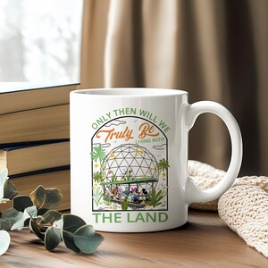 May include: White ceramic mug featuring a colorful illustration of a geodesic dome with cartoon characters and the text "Only Then Will We Truly Be Living With The Land." The mug has a curved handle and is set on a wooden surface.
