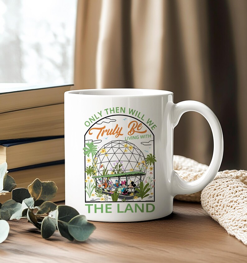 May include: White ceramic mug featuring a colorful illustration of a geodesic dome with cartoon characters and the text "Only Then Will We Truly Be Living With The Land." The mug has a curved handle and is set on a wooden surface.
