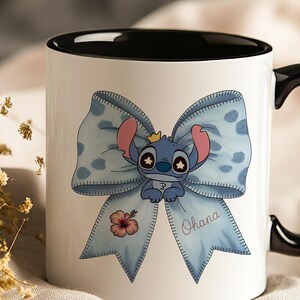 Pu&ograve; includere: Tazza in ceramica bianca con interno e manico neri. La tazza presenta un personaggio dei cartoni animati blu con una corona, su un fiocco azzurro chiaro con pois bianchi. La parola "Ohana" &egrave; scritta sul fiocco, insieme a un fiore rosa.