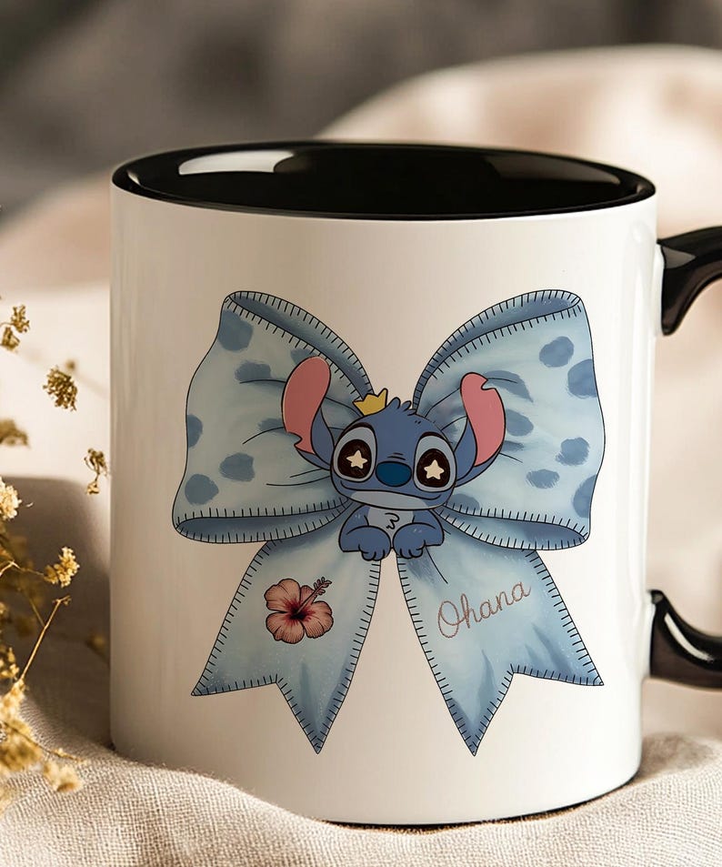 Pu&ograve; includere: Tazza in ceramica bianca con interno e manico neri. La tazza presenta un personaggio dei cartoni animati blu con una corona, su un fiocco azzurro chiaro con pois bianchi. La parola "Ohana" &egrave; scritta sul fiocco, insieme a un fiore rosa.