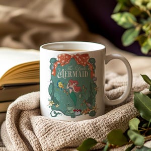 Pode incluir: Caneca de cer&acirc;mica branca com uma ilustra&ccedil;&atilde;o colorida de Ariel de A Pequena Sereia. O design inclui o texto "The Little Mermaid" e um la&ccedil;o vermelho. A caneca est&aacute; sobre uma manta de malha bege.
