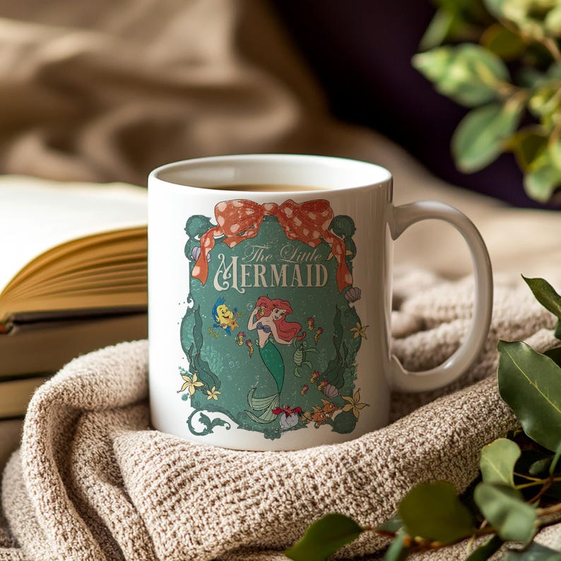 Pode incluir: Caneca de cer&acirc;mica branca com uma ilustra&ccedil;&atilde;o colorida de Ariel de A Pequena Sereia. O design inclui o texto "The Little Mermaid" e um la&ccedil;o vermelho. A caneca est&aacute; sobre uma manta de malha bege.