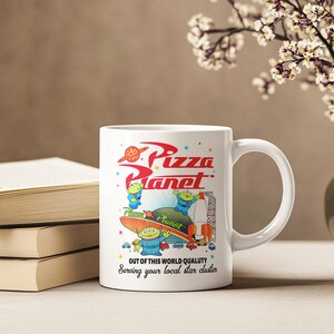 May include: White ceramic mug with a colorful image of the Pizza Planet restaurant from the Toy Story franchise. The image features three alien characters from the movie. The text on the mug reads "Pizza Planet" and "Out of this world quality. Serving your local star cluster."