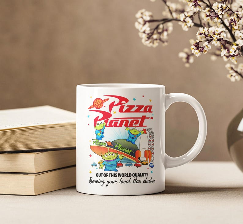 May include: White ceramic mug with a colorful image of the Pizza Planet restaurant from the Toy Story franchise. The image features three alien characters from the movie. The text on the mug reads "Pizza Planet" and "Out of this world quality. Serving your local star cluster."