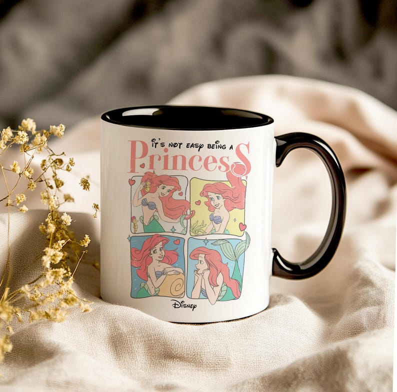 Pode incluir: Caneca de cer&acirc;mica branca com borda e al&ccedil;a pretas. A caneca apresenta uma imagem de desenho animado de Ariel de A Pequena Sereia em quatro poses diferentes. O texto "It's not easy being a Princess" est&aacute; impresso em letras cor-de-rosa na caneca. O logotipo da Disney est&aacute; impresso em preto na parte inferior da caneca.