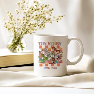 May include: White ceramic mug with a colorful illustration of characters from the movie Toy Story. The mug has the text "Toy Story Est. 1995" printed on it.