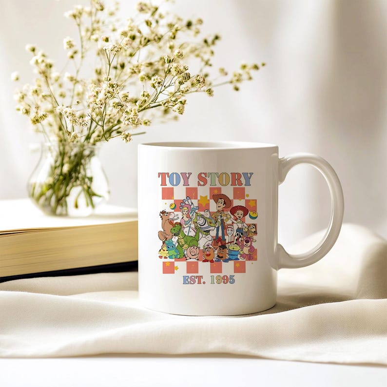 May include: White ceramic mug with a colorful illustration of characters from the movie Toy Story. The mug has the text "Toy Story Est. 1995" printed on it.