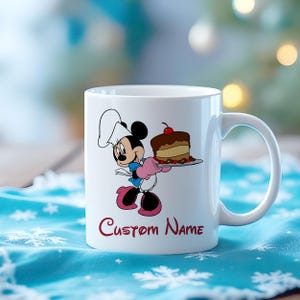 Pu&ograve; includere: Tazza in ceramica bianca con un personaggio dei cartoni animati con un cappello da chef, che tiene in mano una torta su un piatto. La tazza ha un manico curvo e le parole "Custom Name" in corsivo. Il personaggio indossa guanti e scarpe rosa.